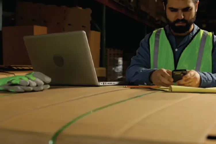 A logistics worker in a warehouse wearing a safety vest, using a smartphone to manage freight bookings.