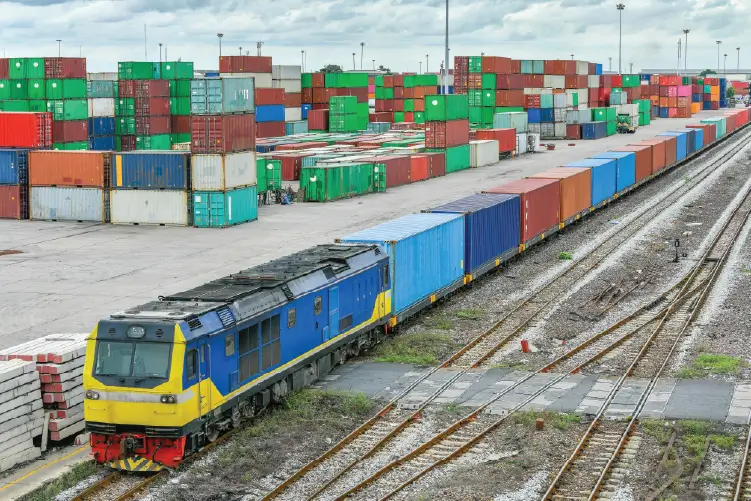 A freight train carrying shipping containers moving through a rail terminal, with a large storage yard filled with stacked containers in the background.