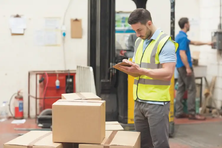 A logistics worker checking inventory on a clipboard in a warehouse, with stacked cardboard boxes and a forklift in the background.