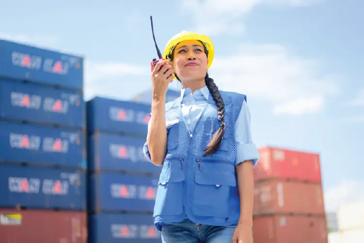 A female logistics worker blue safety vest, communicating via walkie-talkie at a shipping container yard with stacked containers.