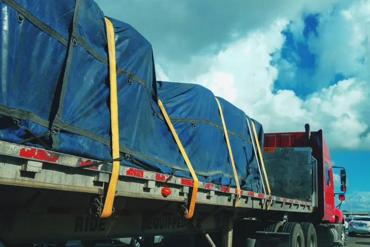 A red truck with a long trailer is carrying cargo covered by a blue tarp, secured with yellow straps, against a cloudy sky.