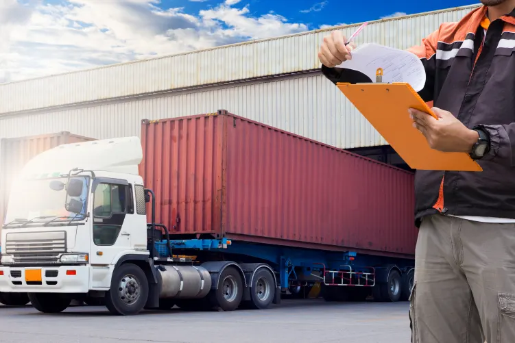 A person wearing a high-visibility jacket holds a clipboard and reviews documents, standing beside a white semi-truck with a large red container in a yard with a building in the background.