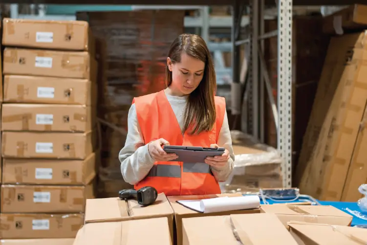 A female warehouse worker using a tablet for inventory management, surrounded by stacked cardboard boxes in a storage facility.