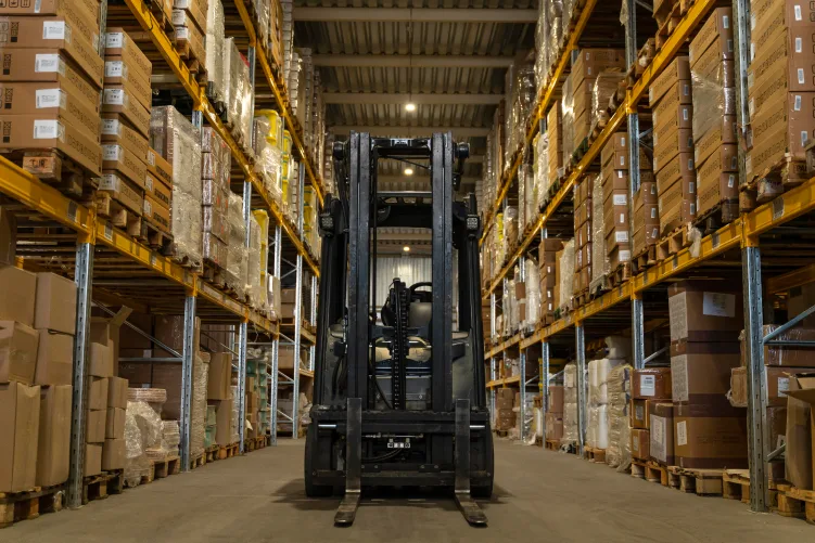 Inside a busy distribution center, showing a forklift facing the camera, positioned between rows of metal shelving filled with various goods and packages.