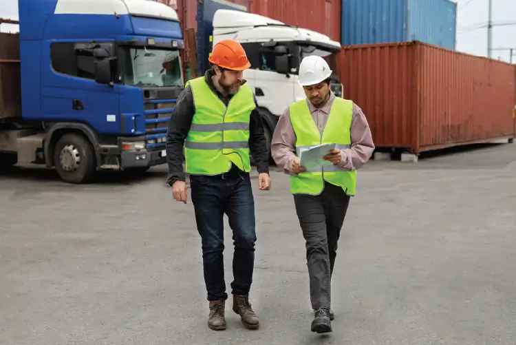 Two logistics professionals discussing freight operations at a shipping yard, with trucks and containers in the background.