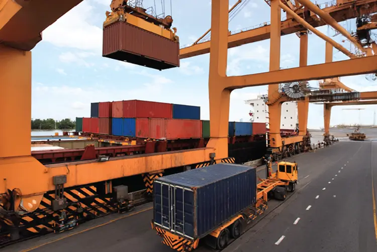 Shipping containers being loaded onto a cargo ship using large cranes at a busy port, with a freight truck transporting a container on the road.