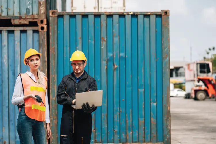 Two logistics professionals in safety gear standing in front of shipping containers, using a laptop to coordinate freight operations.