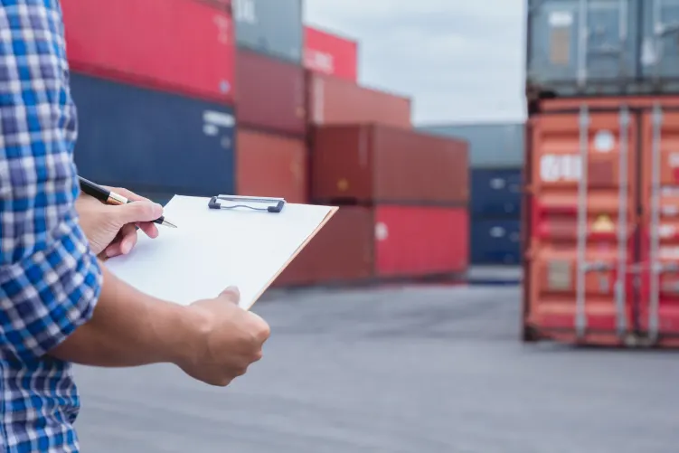 A person in an orange safety vest and hard hat stands near red containers, holding a clipboard and looking at a document. Another person in similar attire is visible in the background.