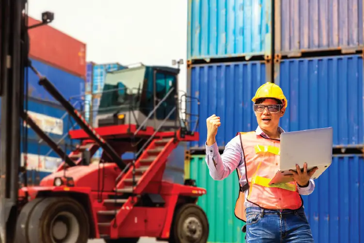 A logistics worker holding a laptop in front of stacked shipping containers