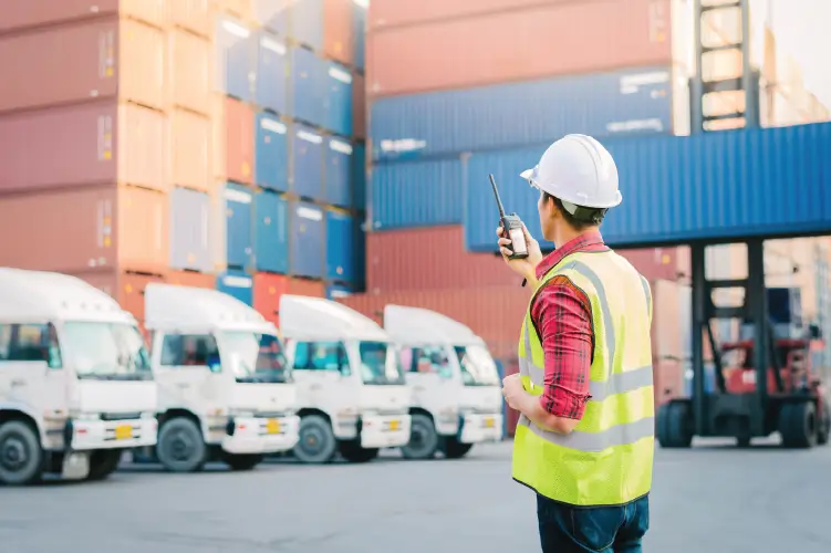 A logistics worker in a safety vest and hard hat using a walkie-talkie to coordinate freight transport, with trucks and stacked shipping containers in the background.