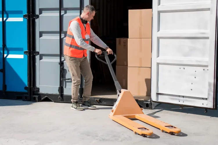 A person in an orange high-visibility vest is guiding a pallet of boxes onto the back of a truck or into a warehouse, with another stack of boxes visible beside them.