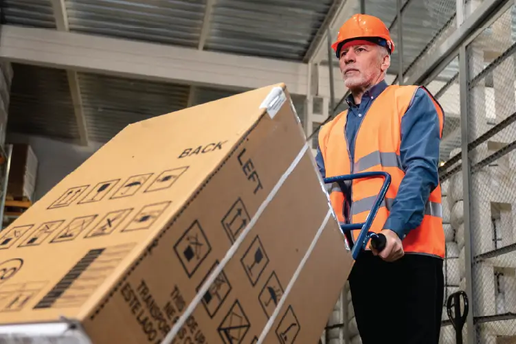A warehouse worker in a safety vest and hard hat carefully transporting a large, labeled cardboard box using a hand truck.