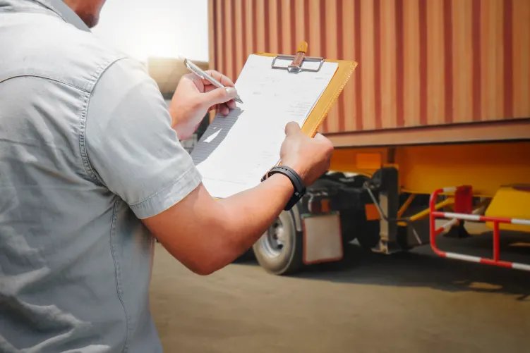 A person seen from behind, holding a clipboard and writing. In the background, part of a truck trailer with an orange container is visible.