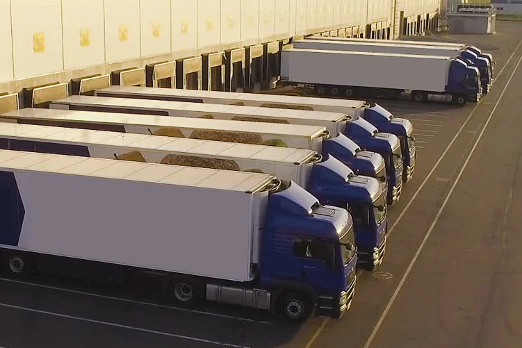An aerial view of multiple semi-trucks with white trailers backed into loading bays at a large freight depot or warehouse, suggesting efficient transport operations.