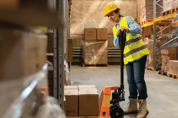 A warehouse worker in a yellow safety vest and hard hat using a pallet jack to move boxes, ensuring safe and secure freight handling.