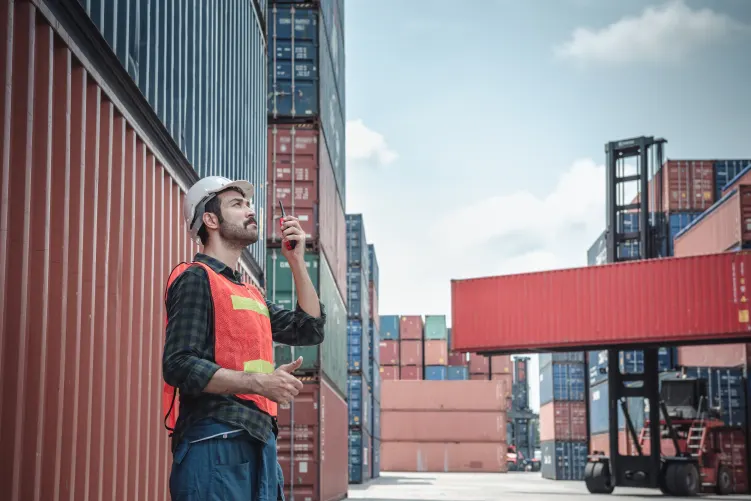 construction or logistics worker in safety gear (hard hats, vests) are looking at a large red shipping container being moved by a crane in an industrial setting.